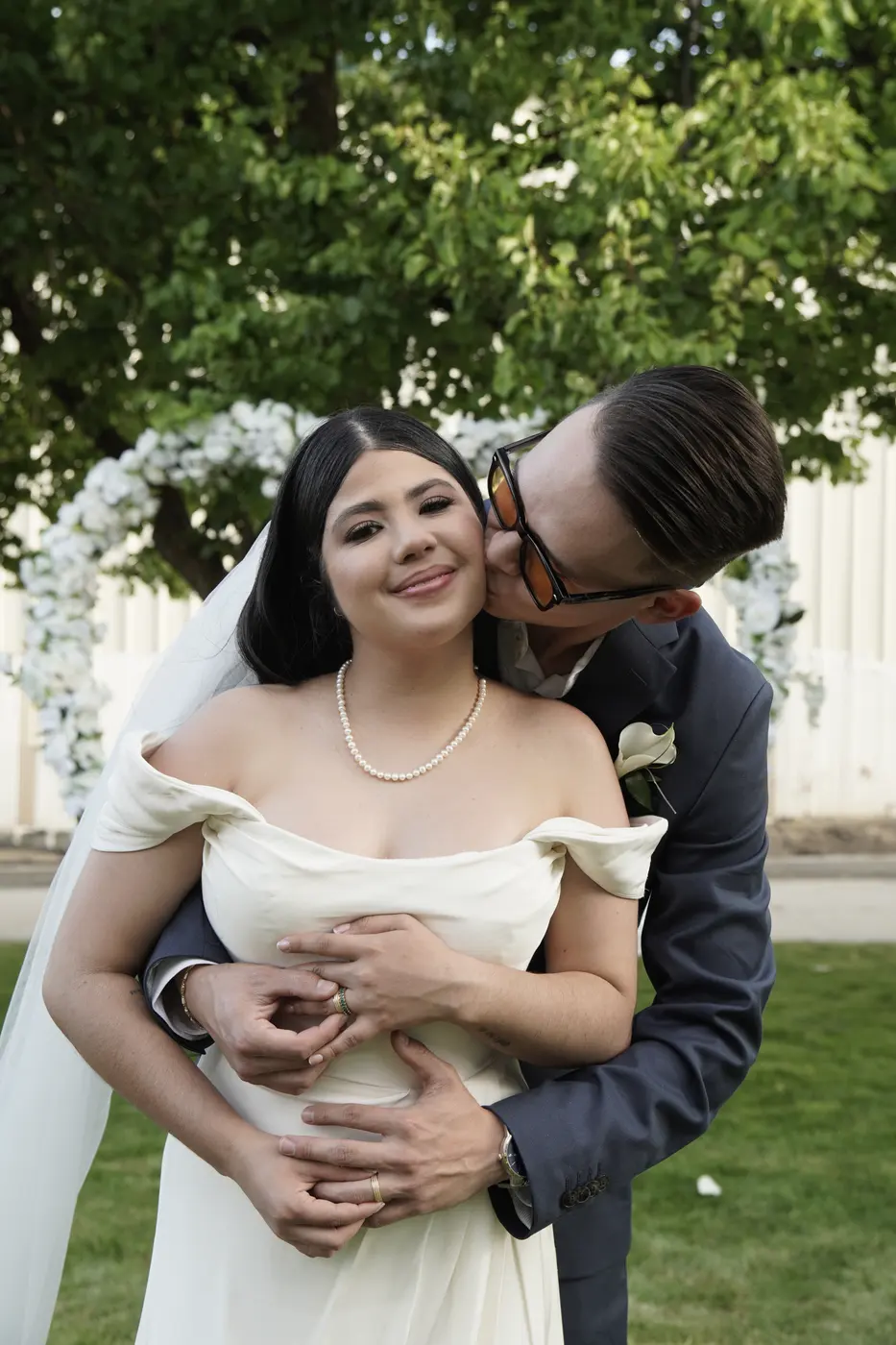 El novio abraza a la novia por la espalda y la besa en la mejilla frente a un arco de flores